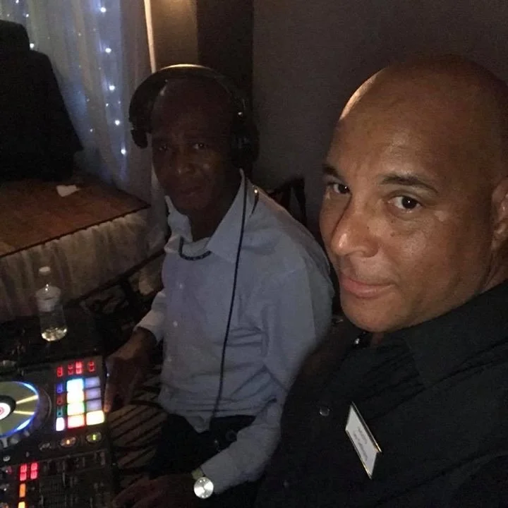 Two men in a dimly lit setting, one wearing a white shirt and headphones, the other wearing a dark shirt with a name tag, standing next to DJ equipment, likely at an event or party.
