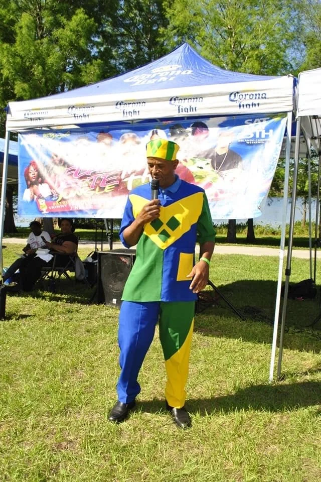 Man dressed in colorful patchwork outfit with a yellow, green, and blue design, wearing a matching hat, standing on grass in front of a tent with a Corona Light banner, holding a microphone during an outdoor event.