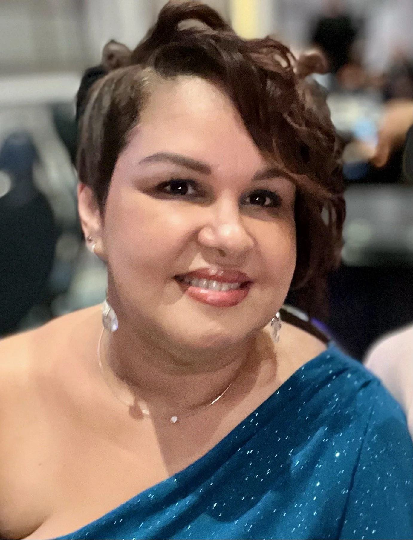 A woman with short curly hair, wearing a blue dress and jewelry, smiling at the camera at a social event.