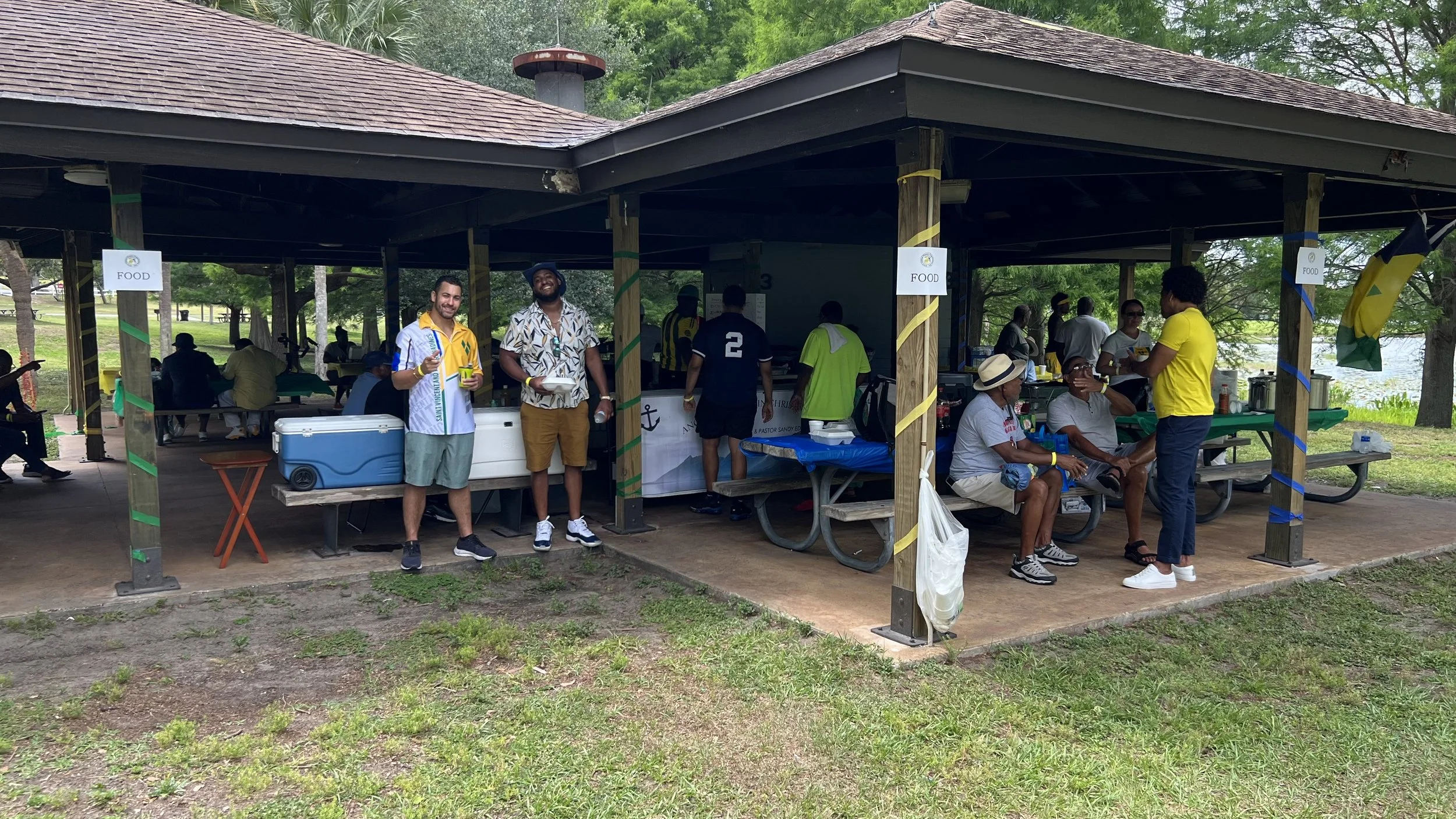 A group of people gathered at a picnic shelter in a park, some sitting at picnic tables and others standing, with food and drinks. The shelter is decorated with yellow, green, and blue ribbons, near a lake and surrounded by trees.