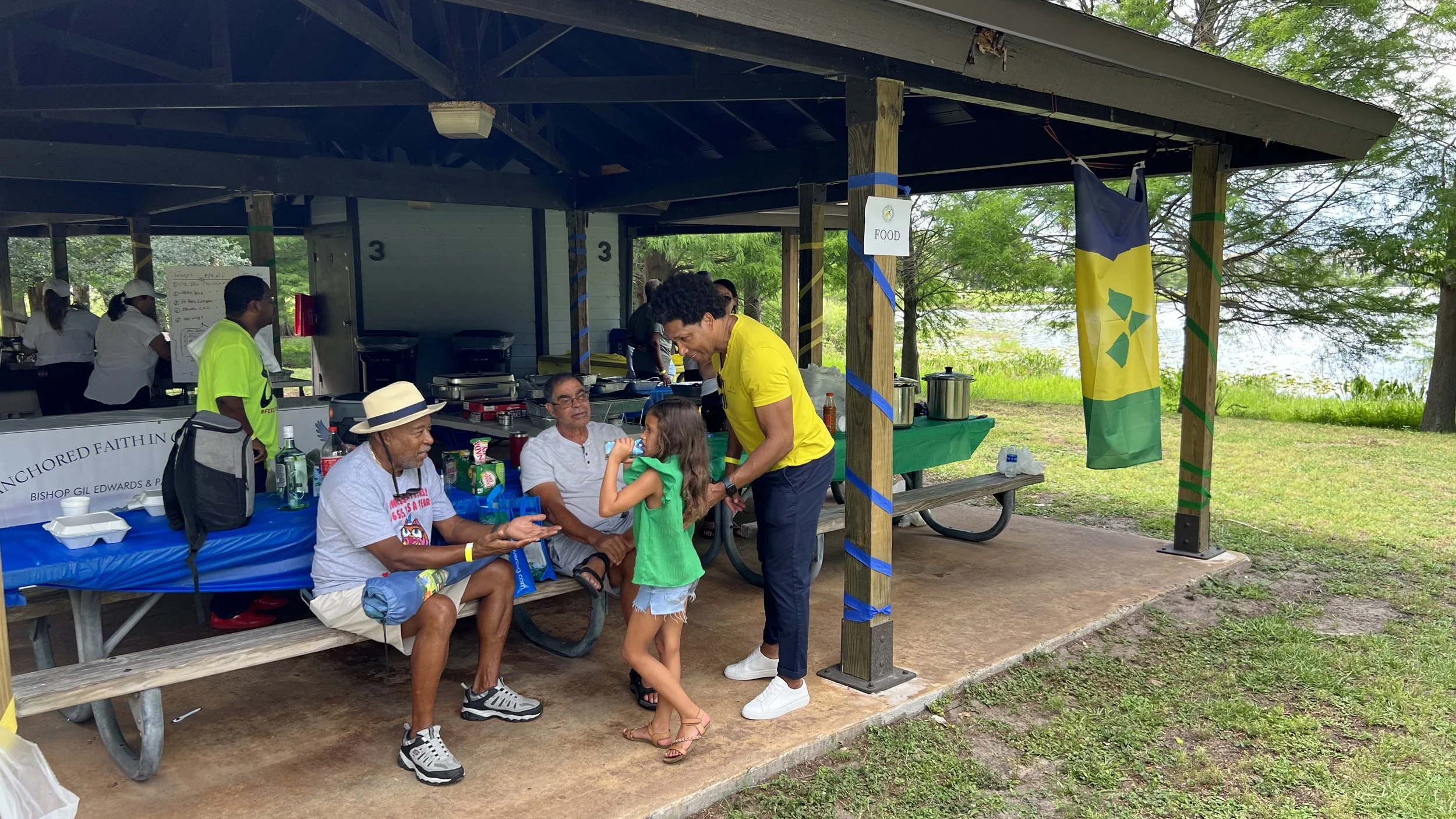 People at an outdoor covered food stand near a lake, with a woman in a yellow shirt talking to a young girl, and two men seated at the table. There are food containers, drinks, and a banner with text and symbols, as well as a flag with green and yell