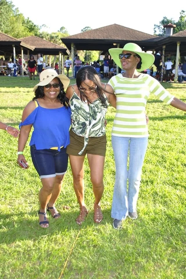 Three women standing outdoors on grass, smiling and holding arms around each other, with a pavilion and people in the background.