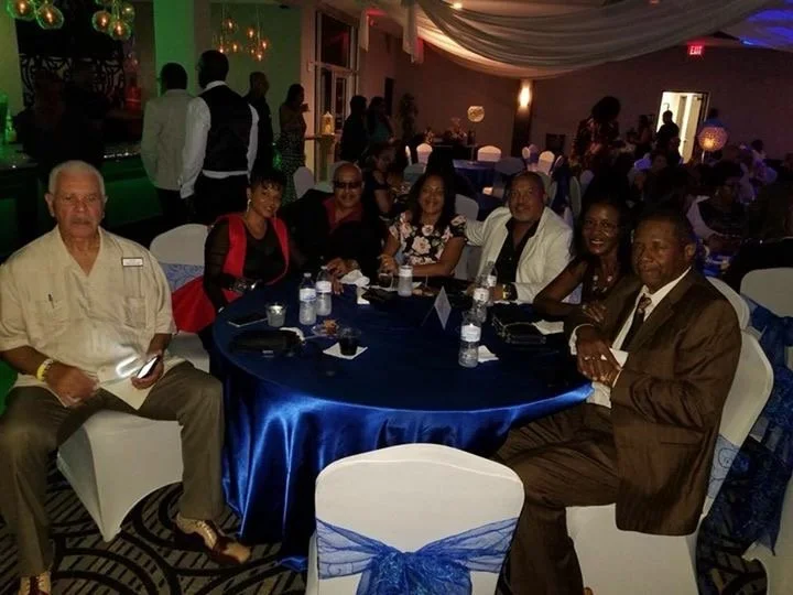 Group of seven people at a banquet table with many water bottles, in a decorated event hall with dim lighting and draped ceiling.