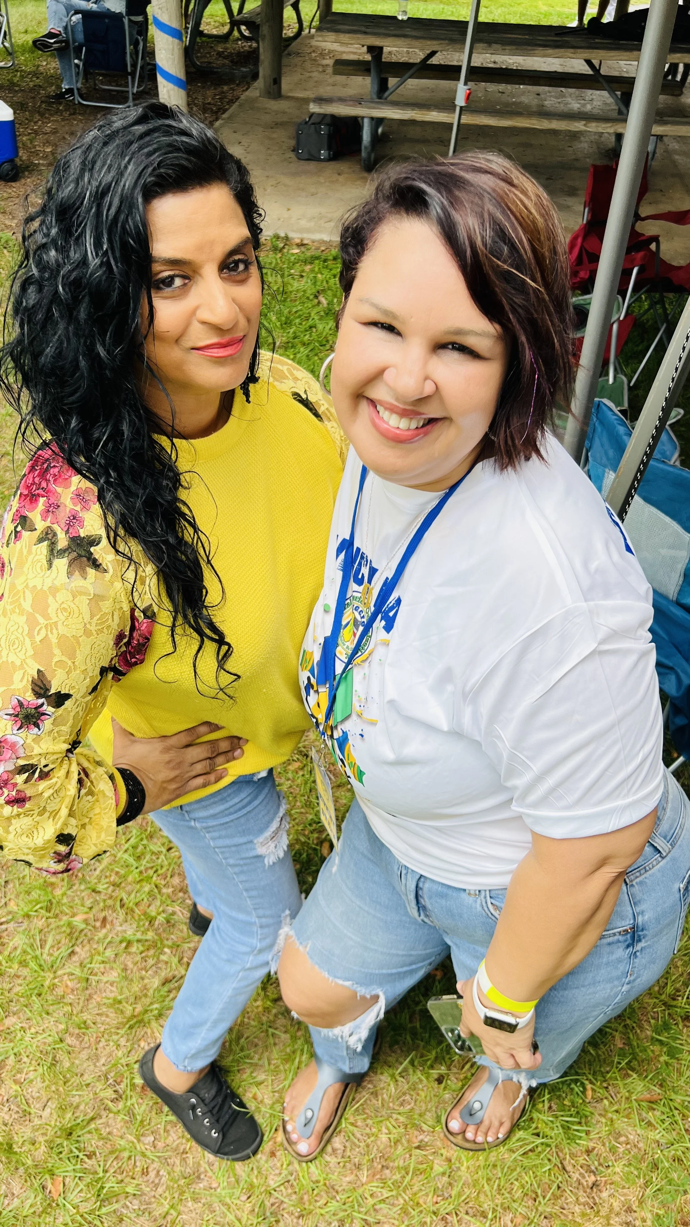 Two women standing outdoors on grass, smiling at the camera. One is wearing a yellow floral dress and the other is in a white T-shirt and ripped jeans. There are picnic tables and chairs in the background.