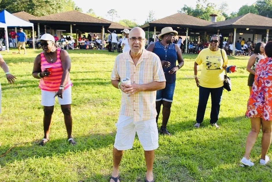 Group of people enjoying an outdoor gathering or party on a sunny day, with a man in the center holding a drink and others socializing on a grassy area with pavilion structures in the background.