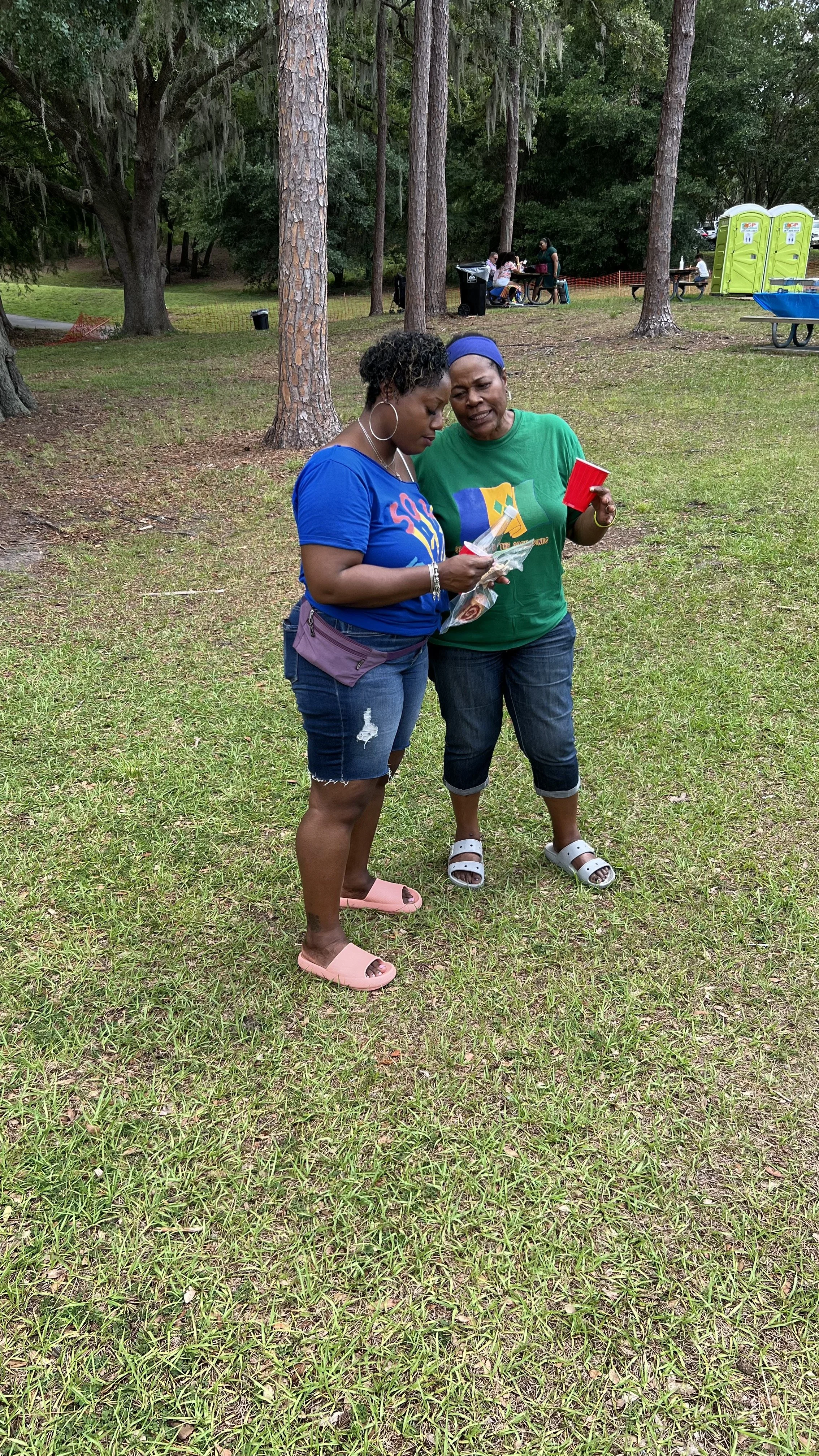 Two women standing outdoors on grass, looking at something in their hands. One is wearing a blue shirt, denim shorts, and pink sandals, while the other is dressed in a green shirt, black capris, and white sandals. Trees and portable toilets are visib