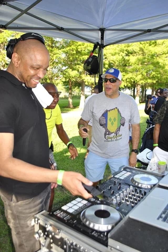 A group of people at an outdoor event, with one person using DJ equipment under a canopy, others smiling and talking in a park setting with trees in the background.