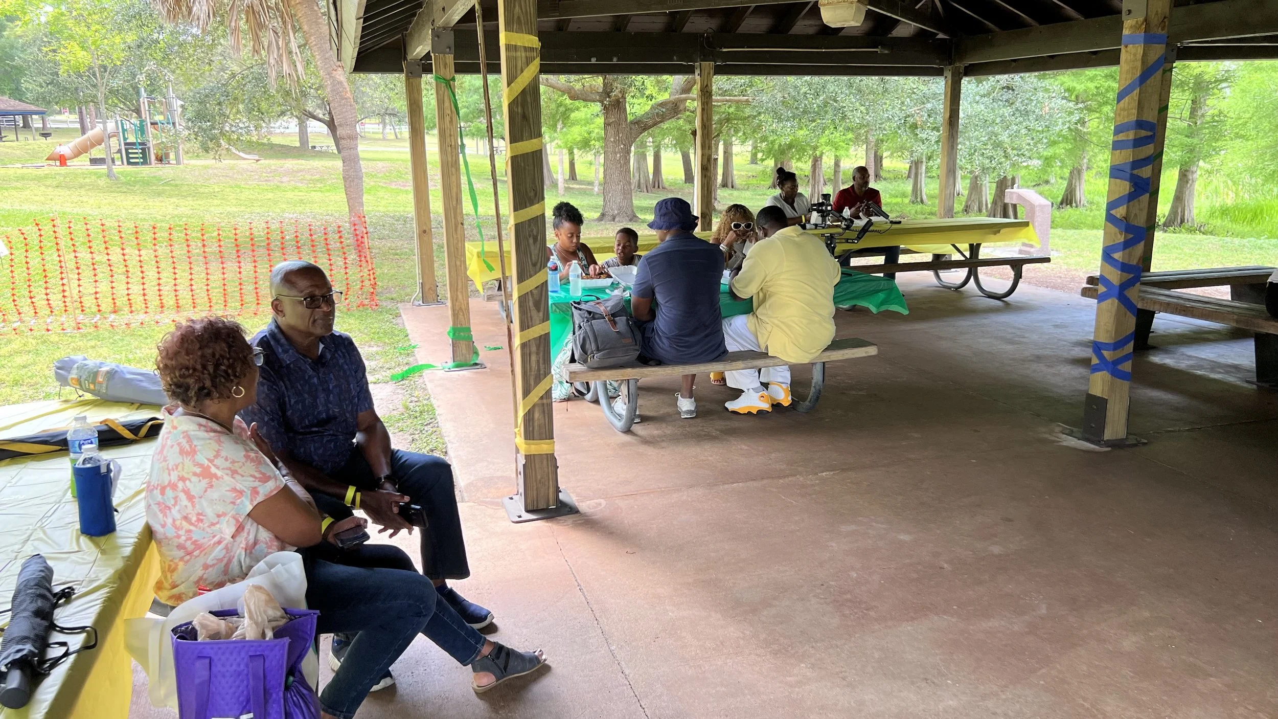 People sitting and talking at picnic tables under a pavilion in a park with trees and a playground in the background.