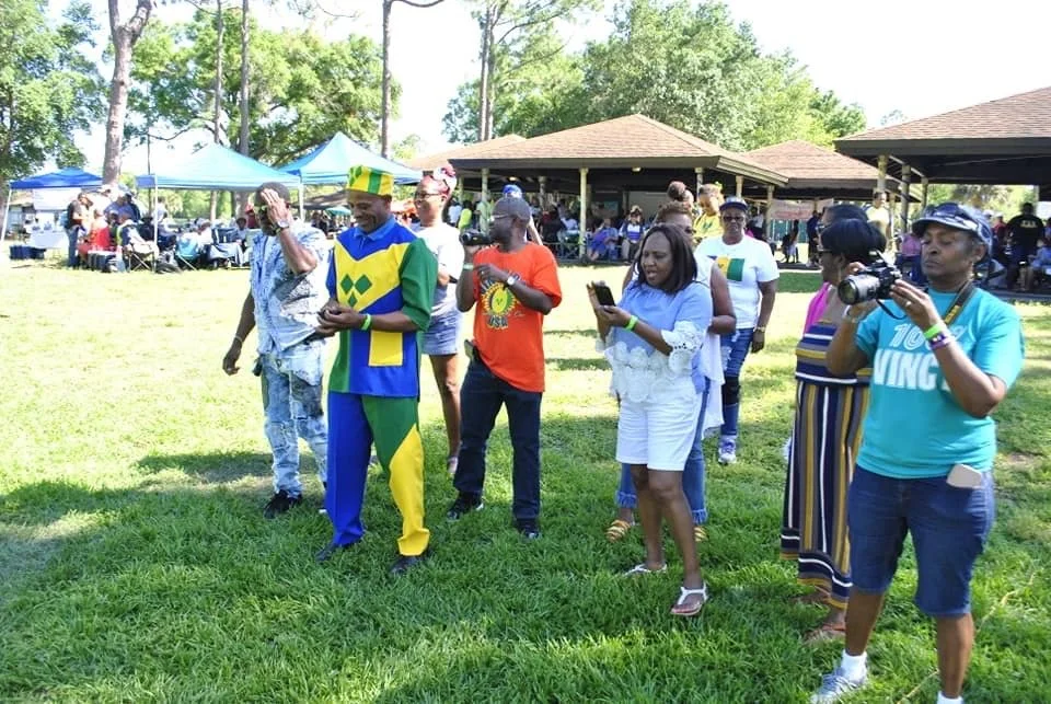 Group of people standing on grass at outdoor event, some taking photos or videos, with tents and pavilion in background.