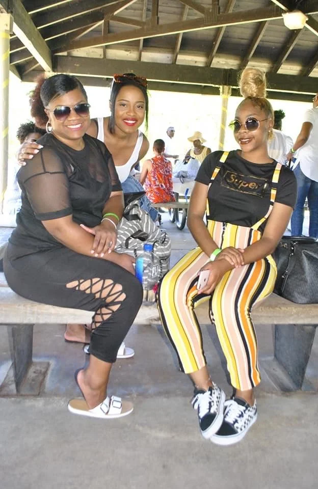 Three women sitting on a picnic table under a pavilion, smiling and enjoying each other's company; one wearing black, another in white and striped pants, and the third in a black top with yellow striped pants.