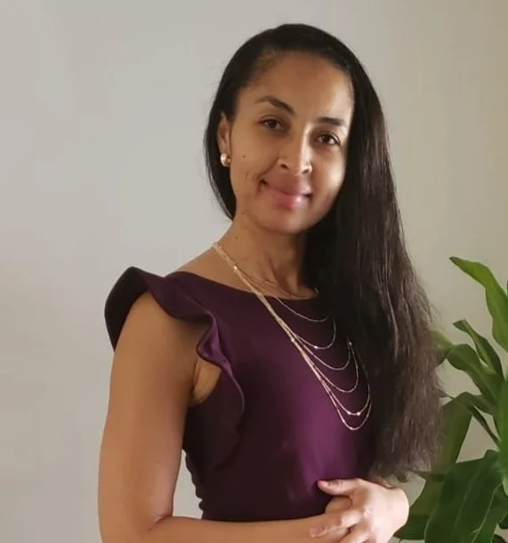 Woman with long dark hair, wearing a purple dress with ruffled sleeves, gold jewelry, standing next to a green plant.
