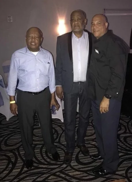 Three men standing together indoors, dressed in formal attire, with patterned carpet and a wall with a light fixture in the background.