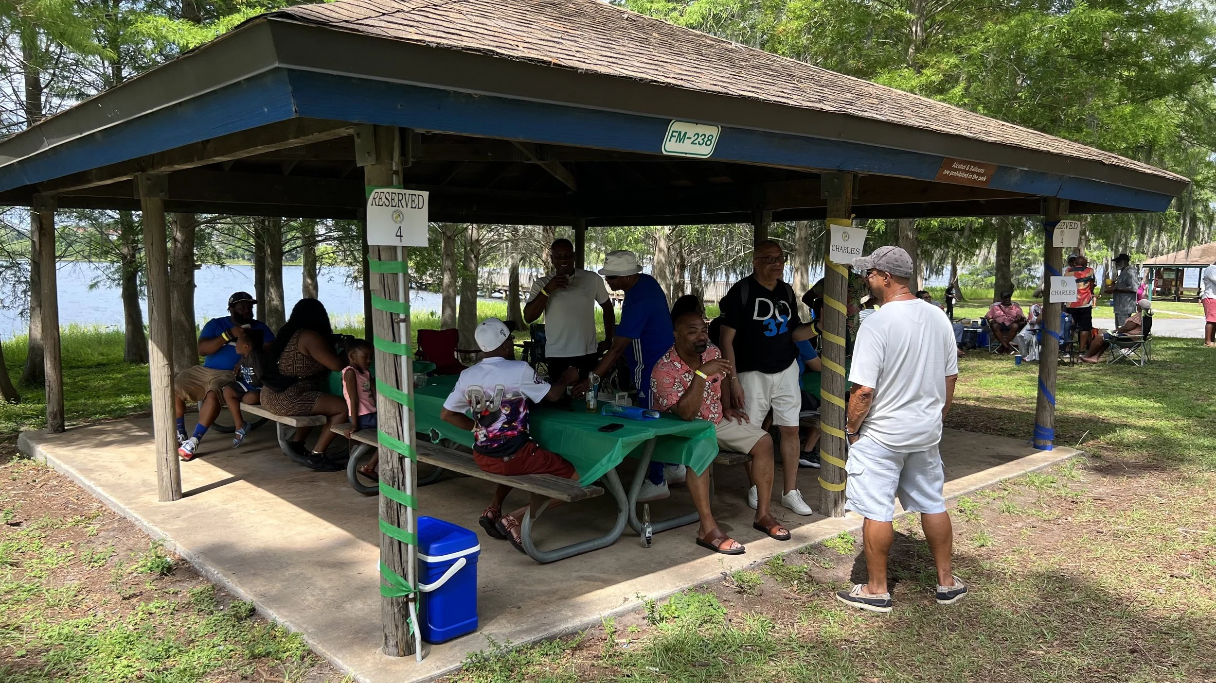 A group of people gathered under a wooden pavilion near a lake in a park, sitting and standing around tables, some engaging in conversation, with trees and other pavilions visible in the background.
