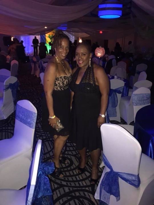 Two women in elegant black dresses standing in a decorated banquet hall with rows of chairs and blue ribbon accents.