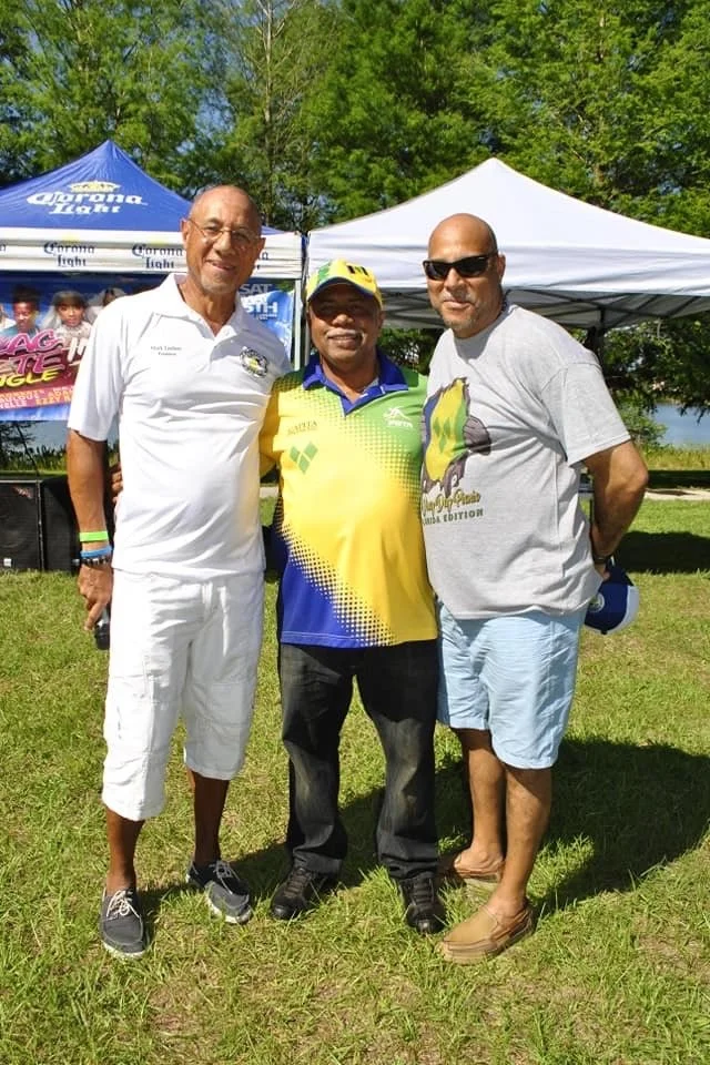 Three men standing outdoors on grass, smiling, with tents and trees in the background, at a sunny event.