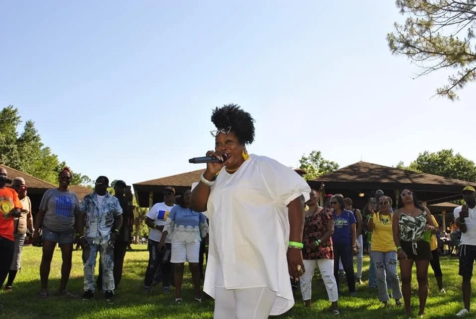 A woman in white speaking into a microphone at an outdoor gathering, with a group of people standing behind her on a grassy area under a clear sky.