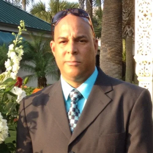 A man in a dark suit, light blue shirt, and patterned tie standing outdoors with palm trees, lush green plants, and a building in the background.
