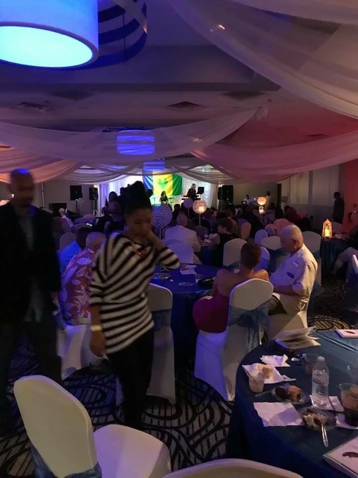 People gathered at a banquet hall or event space with decorated ceilings, dining tables, and a stage with musicians or performers performing in front of a South African flag.