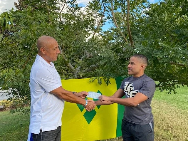 An older man and a younger man shaking hands during a card handover in front of a yellow flag with a green emblem, outdoors with trees and grass.
