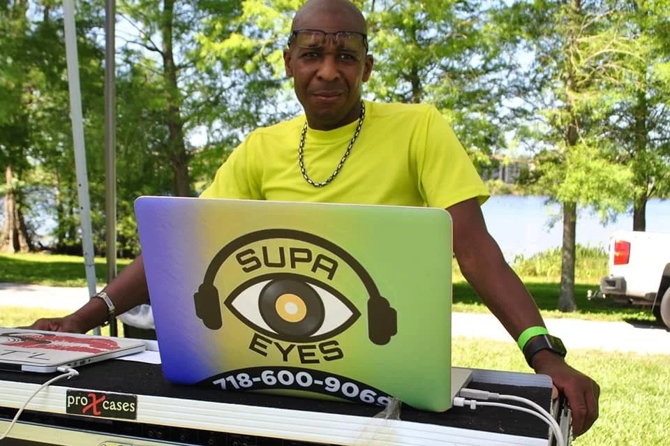 A man wearing a bright yellow shirt and sunglasses standing outdoors behind a DJ setup with a laptop displaying a logo that reads 'SUPA EYES' with an eye and headphone design.