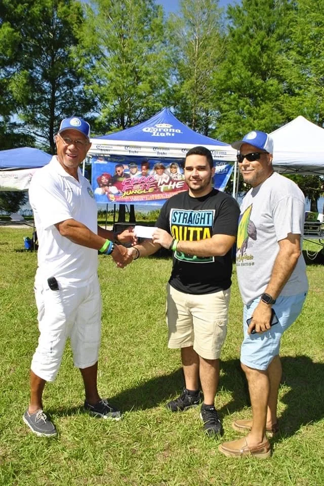 Three men standing outdoors on grass, shaking hands, with a blue tent and trees in the background. One man is wearing a white outfit and cap, the second man is in a black T-shirt and beige shorts, and the third is in a gray T-shirt and light blue shorts.