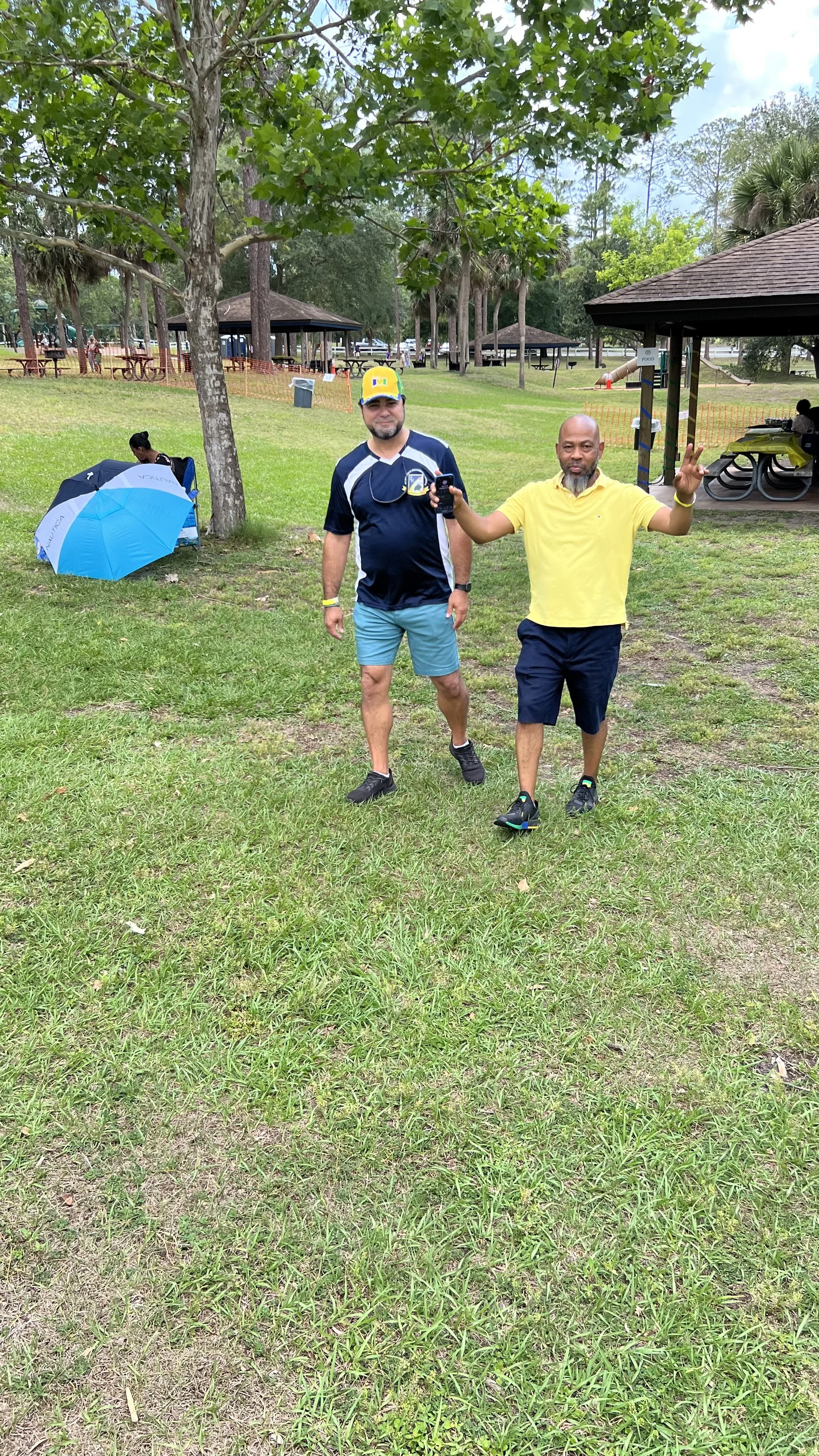 Two men walking on a grassy park area, one holding a phone. One man is wearing a blue and white sports outfit with a yellow cap, and the other man is wearing a yellow shirt and shorts. In the background, picnic shelters, trees, a woman sitting under 