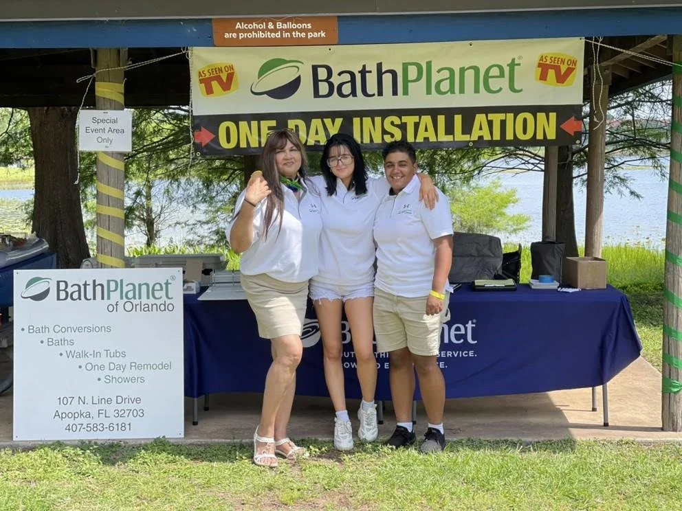Three women standing together in front of a promotional booth for Bath Planet, posing for a photo with the river in the background. The booth promotes a one-day installation event.