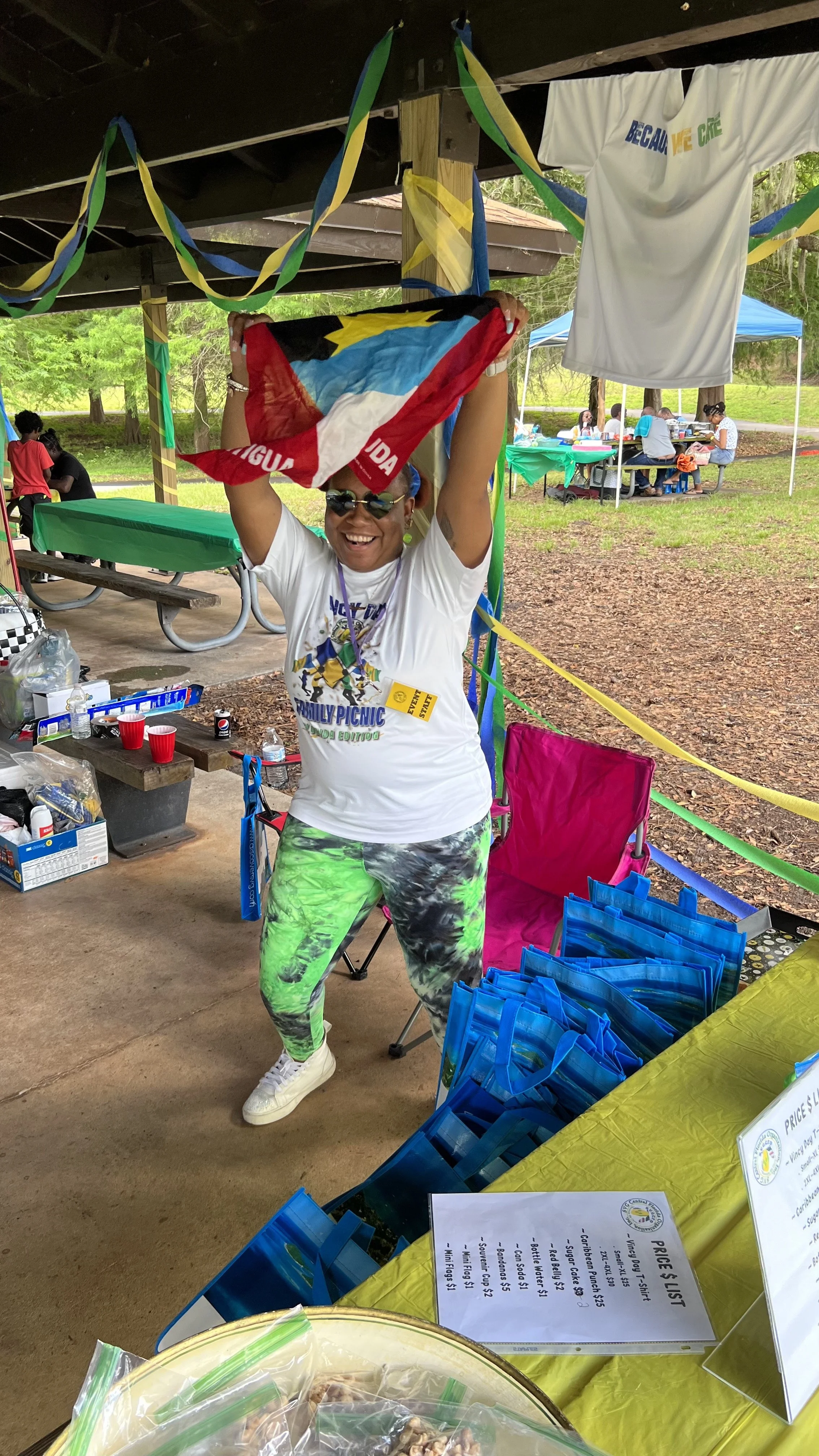 Smile woman at a picnic, holding up a flag, wearing sunglasses and colorful leggings, surrounded by tents and picnic tables.