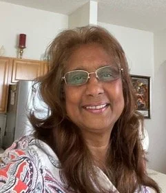 A woman with long, wavy brown hair, glasses, and a patterned top, smiling in a room with a wooden cabinet and wall art.