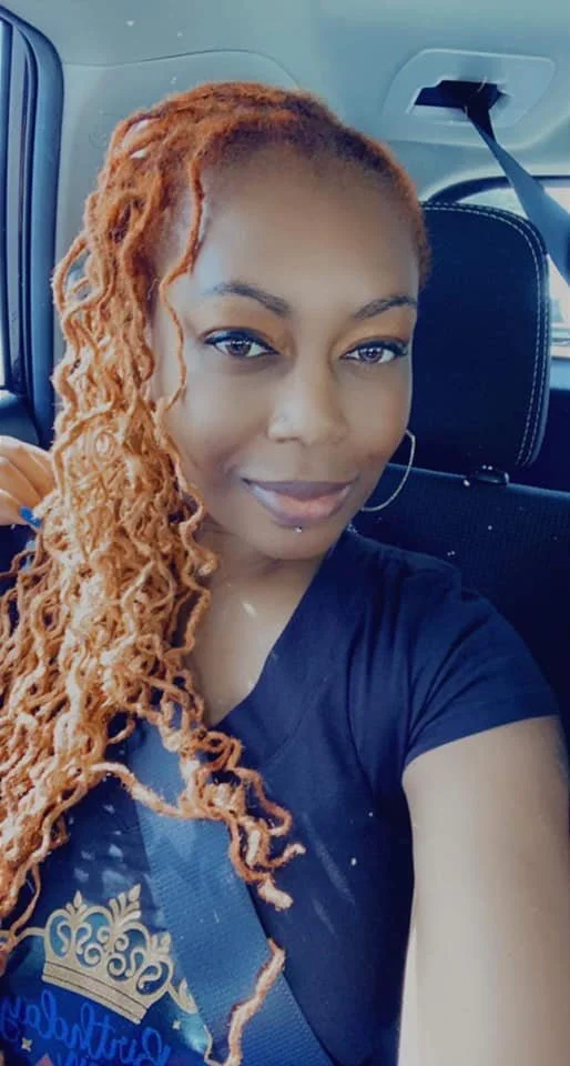 A woman with long, curly, reddish-blonde hair taking a selfie in a car. She is smiling slightly, wearing a black top, large hoop earrings, and a seatbelt.