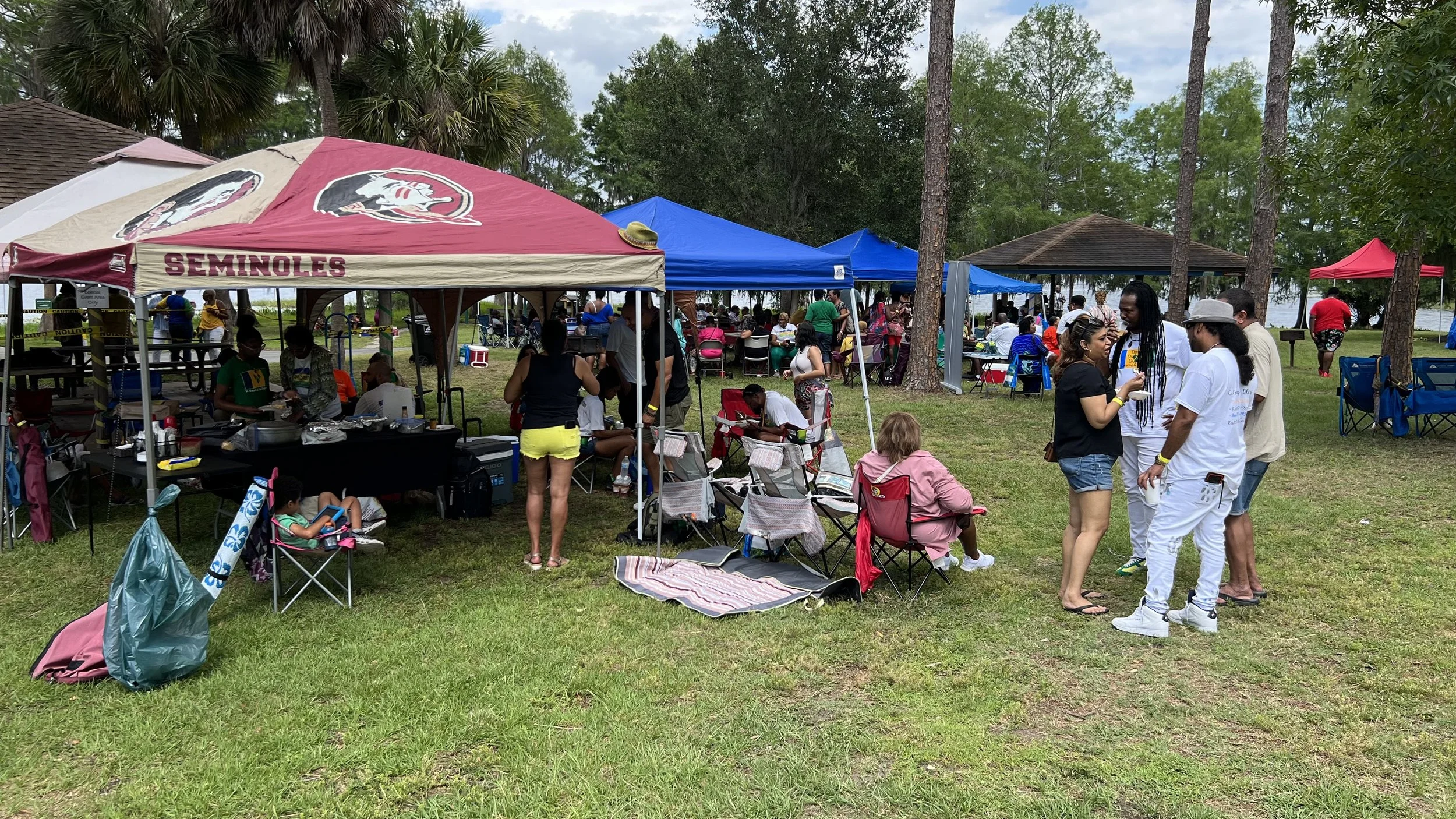 People gathered outdoors around tents and tables at a park or lakeside area, socializing and relaxing under trees with a lake visible in the background.