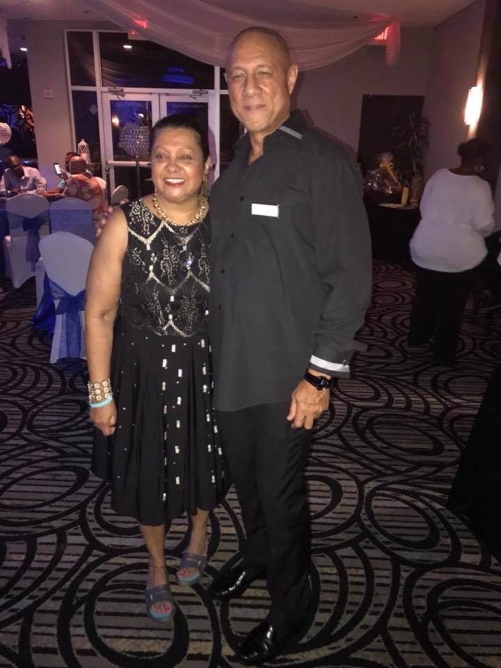 A woman and a man dressed formally, standing together in a banquet hall decorated for an event, smiling at the camera.