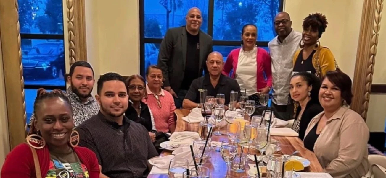 A group of people at a restaurant table, smiling, with some seated and others standing behind. The table has drinks and glasses, and the background shows windows with a blue sky outside.