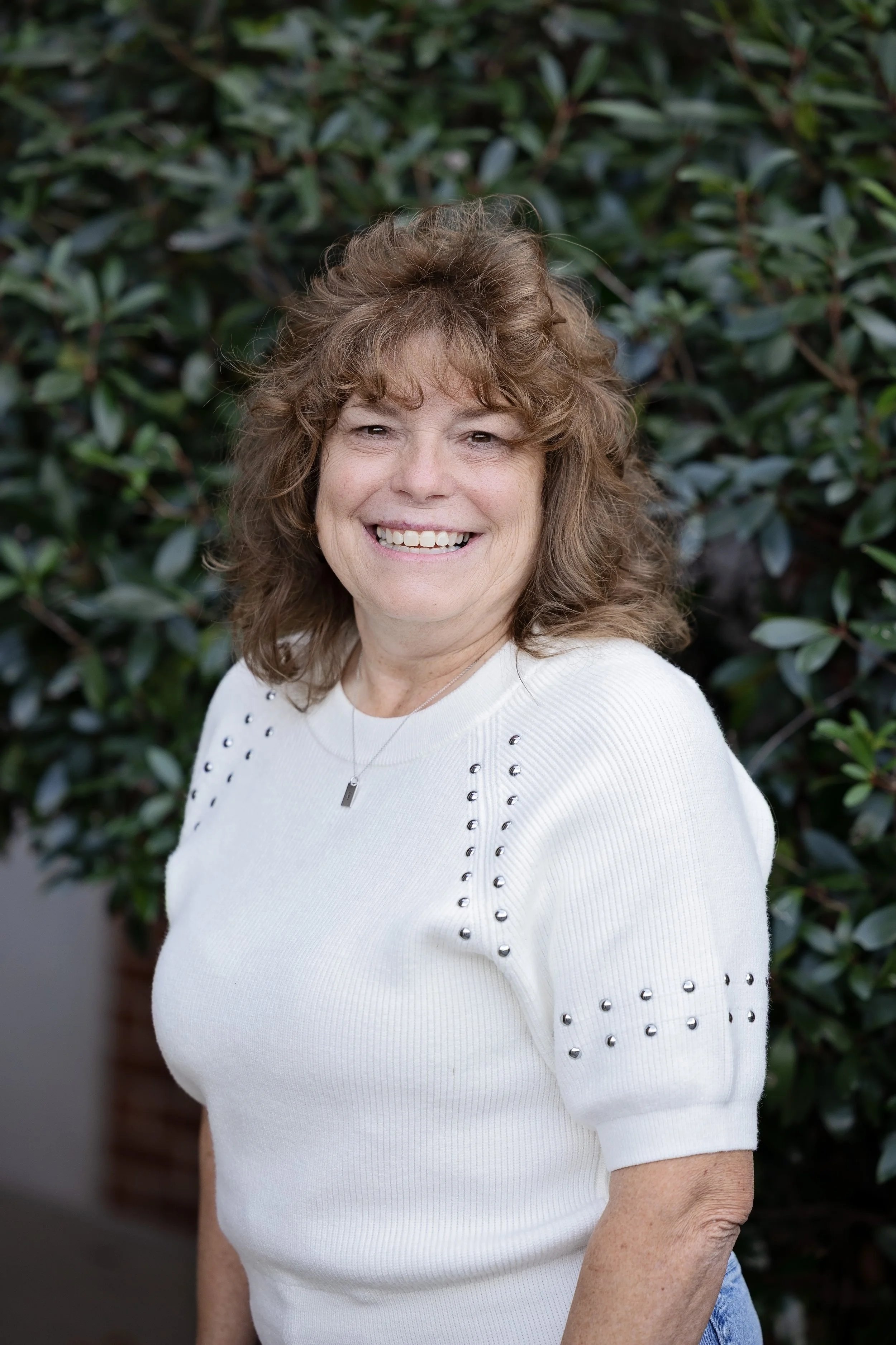 Smiling woman with curly hair wearing a beige knitted sweater against a light gray background.
