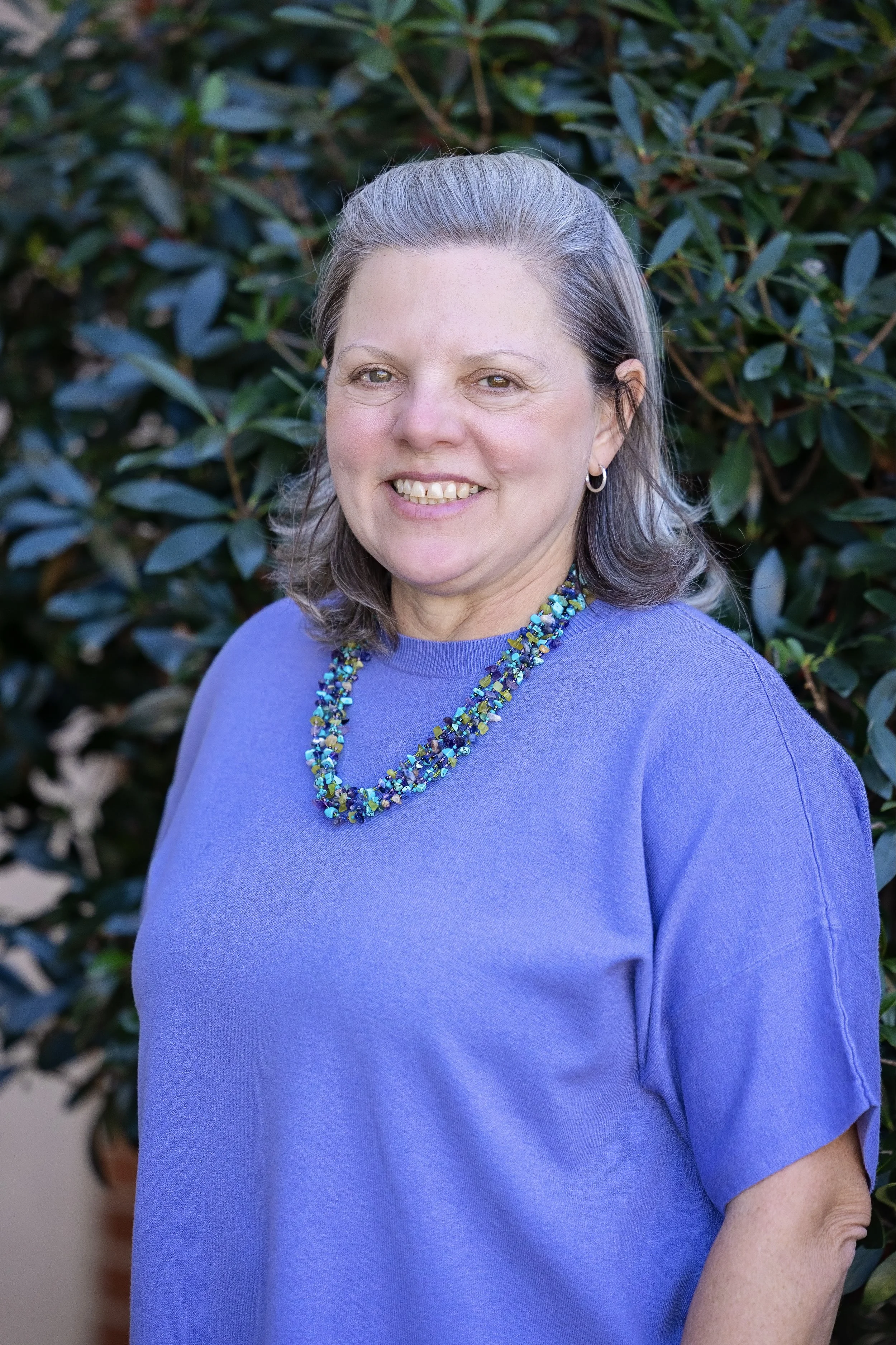 Woman with short gray hair wearing a blue shirt and a silver cross necklace, smiling against a light gray background.