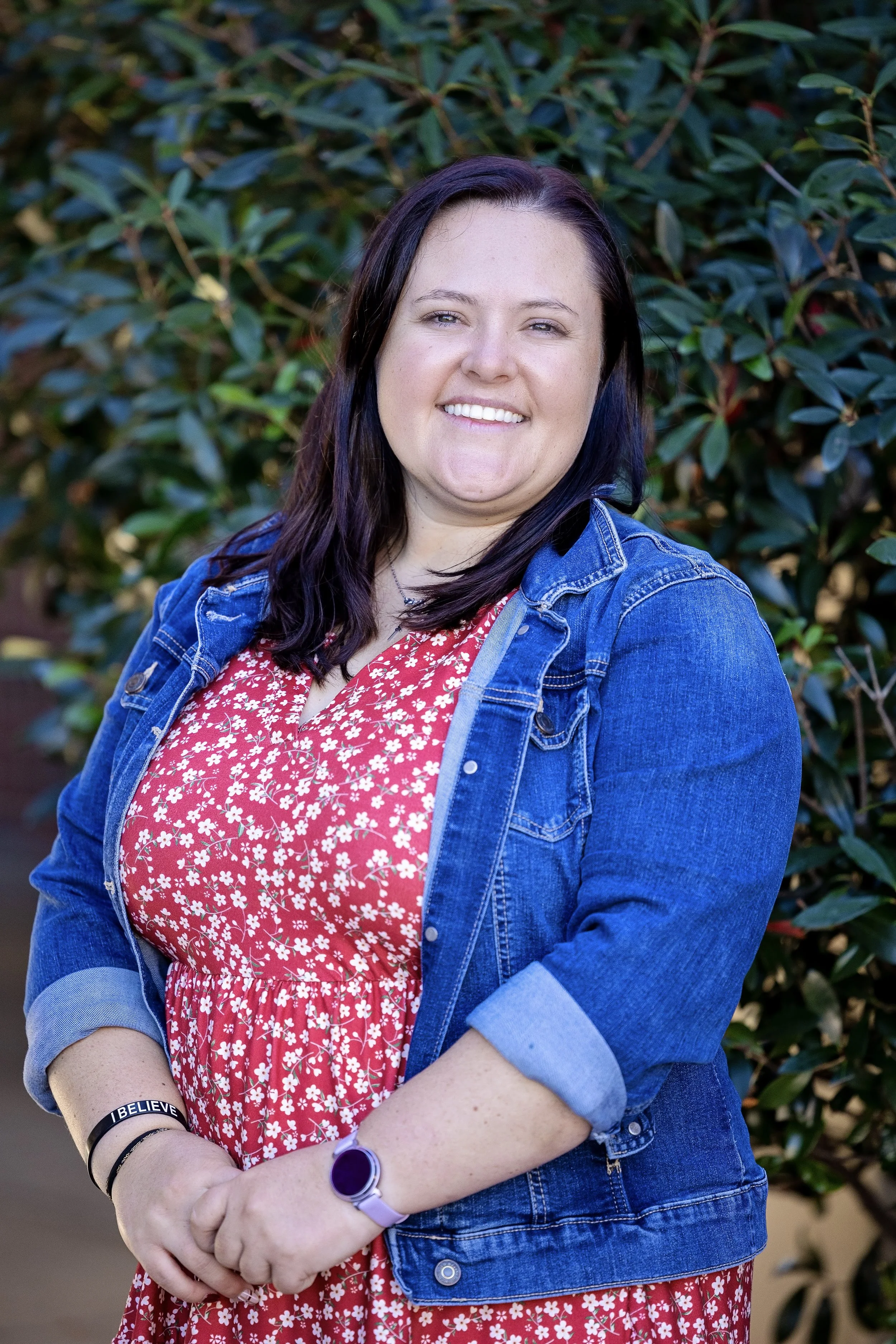 Woman smiling, wearing a black vest and red sweater, pendant necklace, with long dark hair, against a gray background.