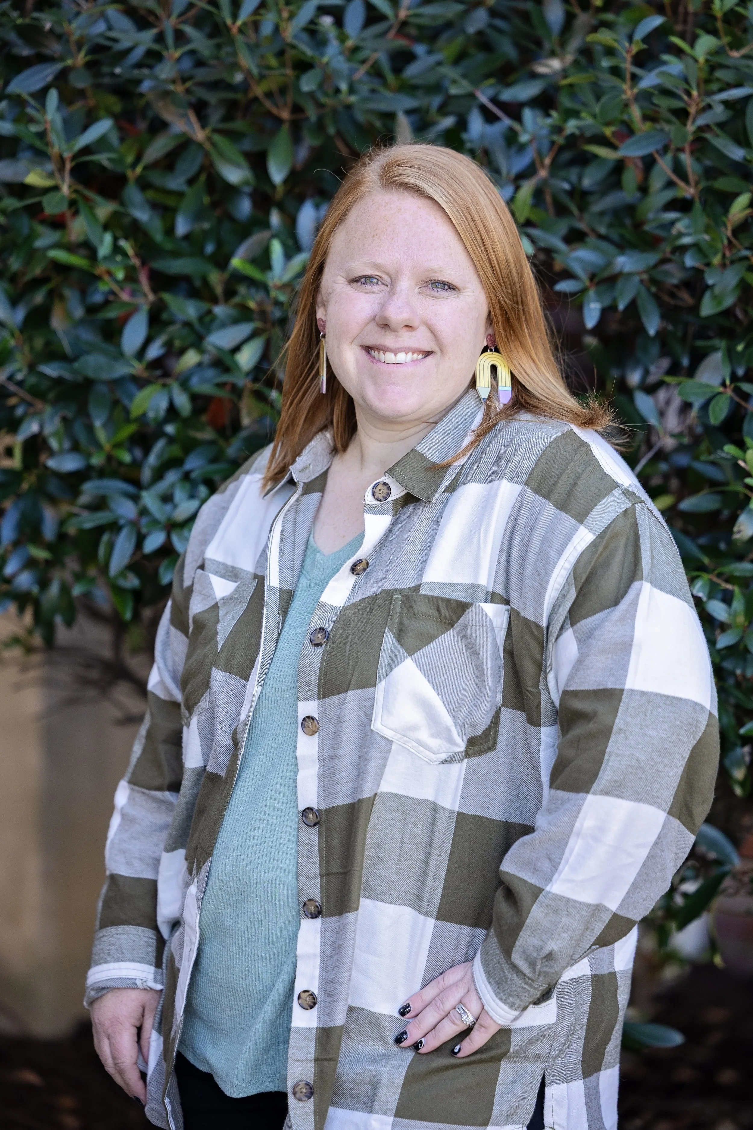 Smiling person with red hair, wearing a dark blue top and circular earrings, against a light gray background.