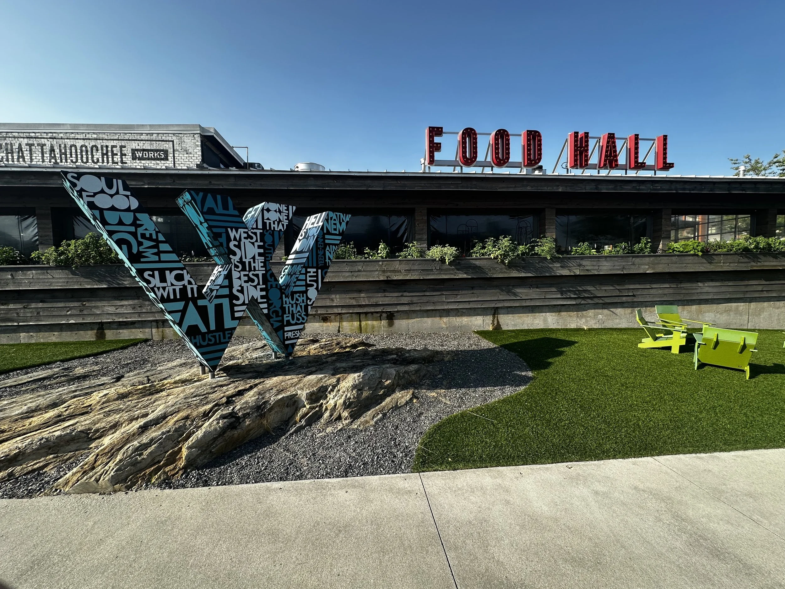 Painted steel “W” logo sculpture at The Works ATL in Atlanta—black base coat with hand-lettered Southern dialect typography. Public art by John Tindel.