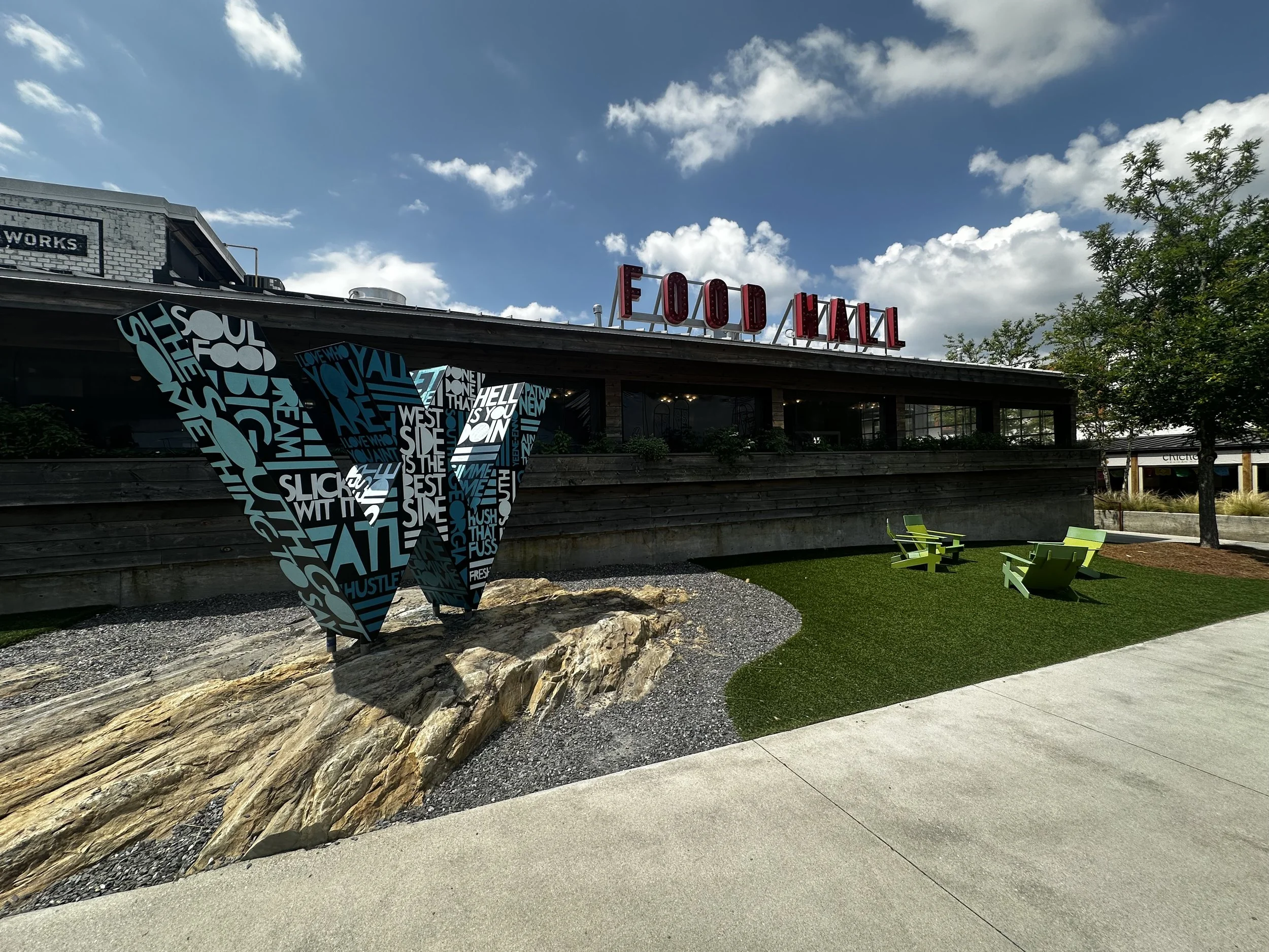 Painted steel “W” logo sculpture at The Works ATL in Atlanta—black base coat with hand-lettered Southern dialect typography. Public art by John Tindel.