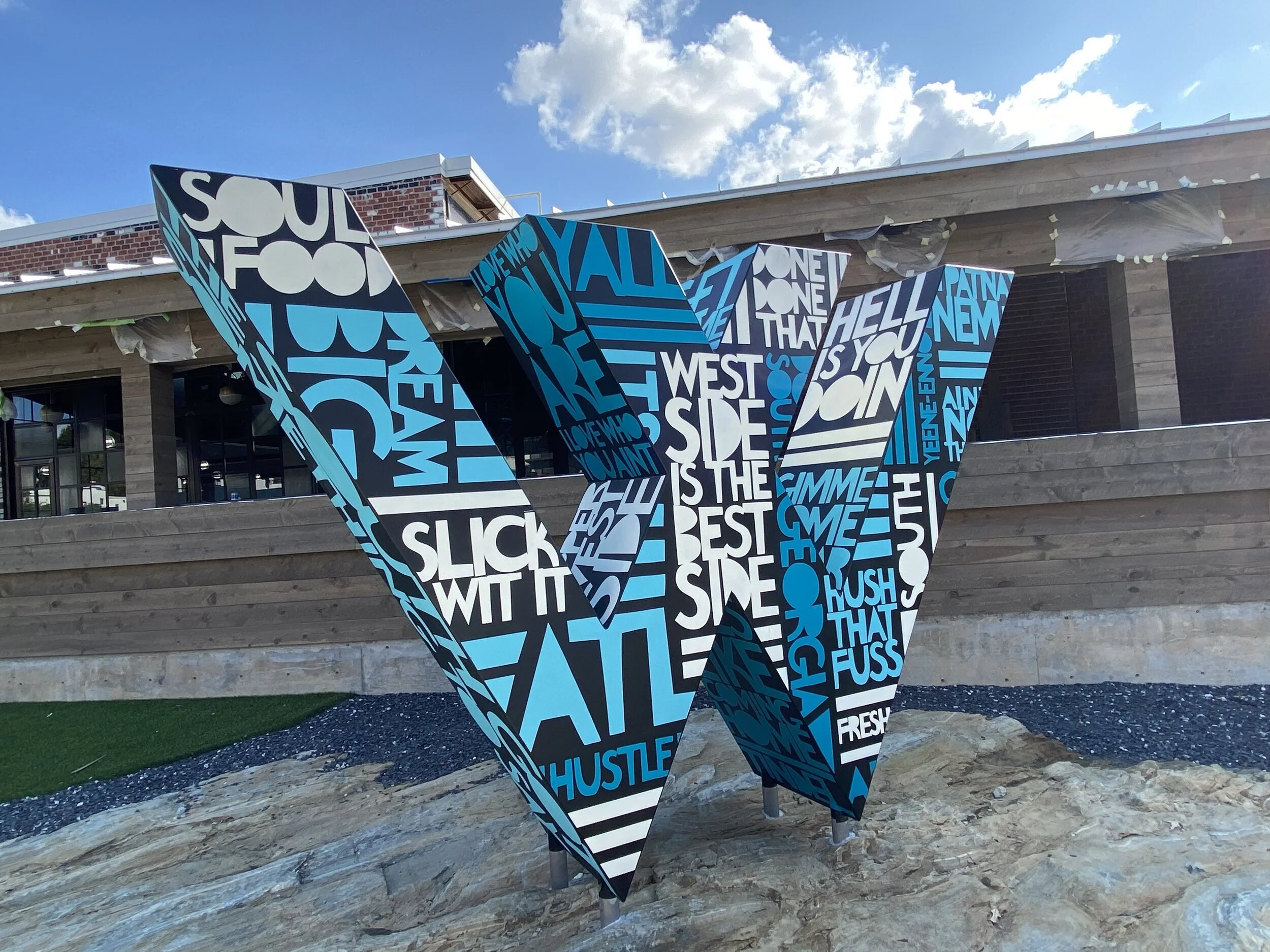 Painted steel “W” logo sculpture at The Works ATL in Atlanta—black base coat with hand-lettered Southern dialect typography. Public art by John Tindel.