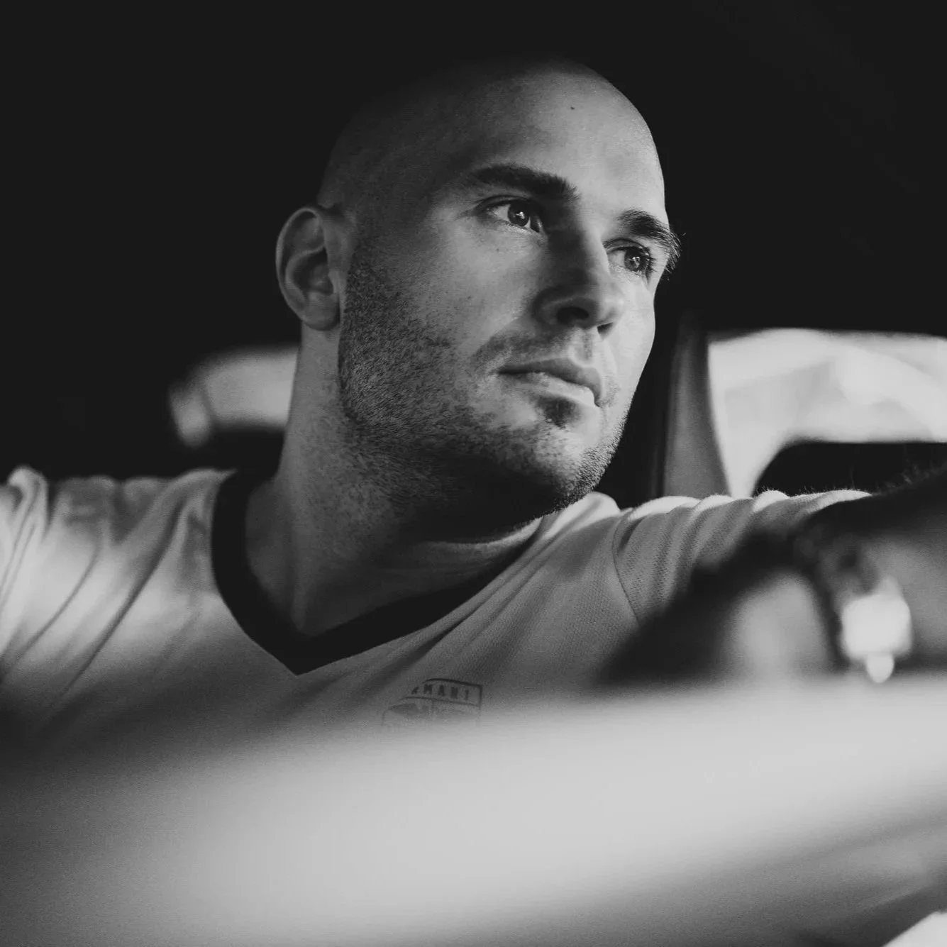 A young man with a buzz cut and stubble looking thoughtfully through a vehicle window in black and white.