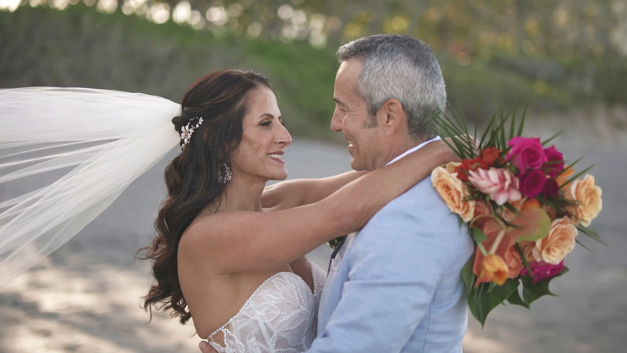A bride and groom smiling at each other, holding each other close on their wedding day outdoors with a natural background. The bride is wearing a lace wedding dress and veil, and is holding a bouquet of flowers behind the groom's back.