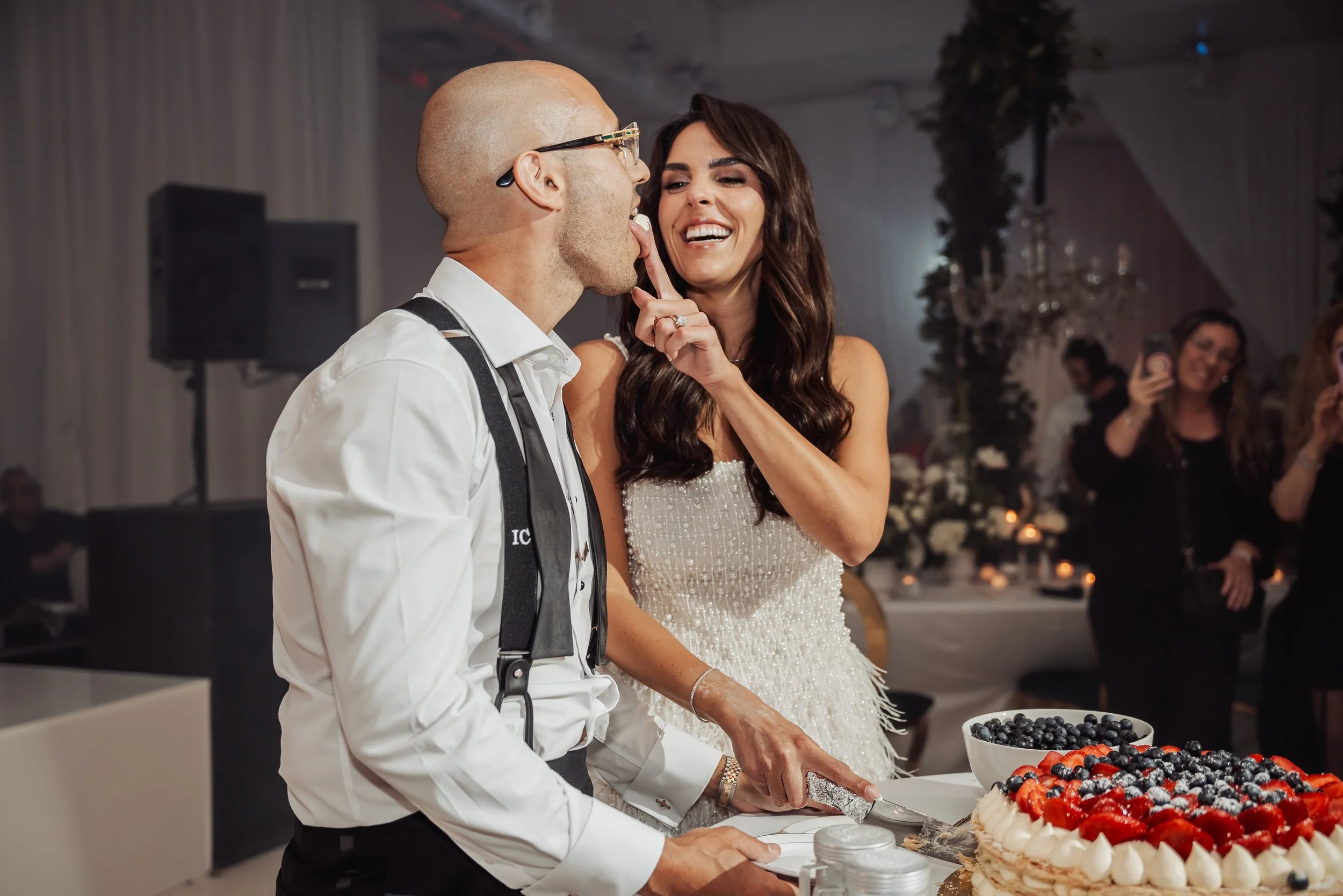 A couple at a wedding reception, with the woman playfully touching the man's tongue, while he is about to cut a cake decorated with berries.