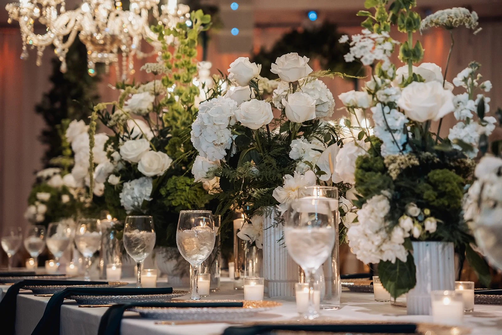 Elegant table centerpiece with large white floral arrangements including roses and hydrangeas, surrounded by candles and glassware at a formal event.