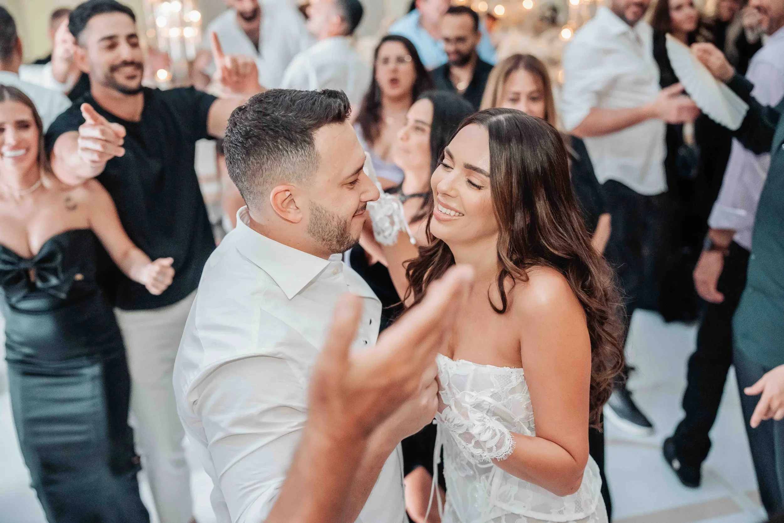 Couple dancing closely at a lively celebration, surrounded by smiling guests in elegant attire.