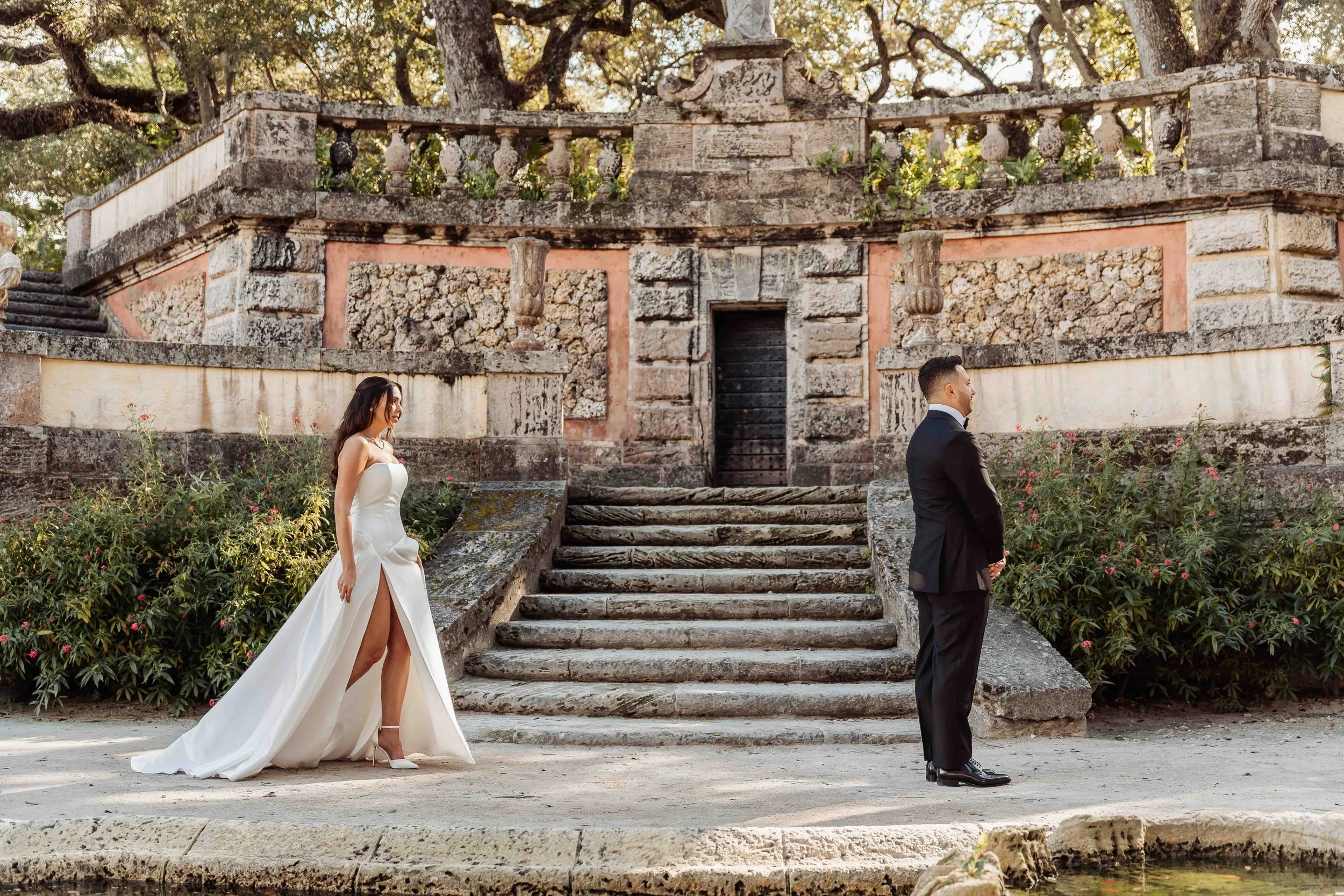 A bride and groom stand apart facing away from each other outdoors with an old stone staircase and ornate stone railing behind them. The bride is in a white wedding gown with a slit and high heels, and the groom is in a black tuxedo, both looking in