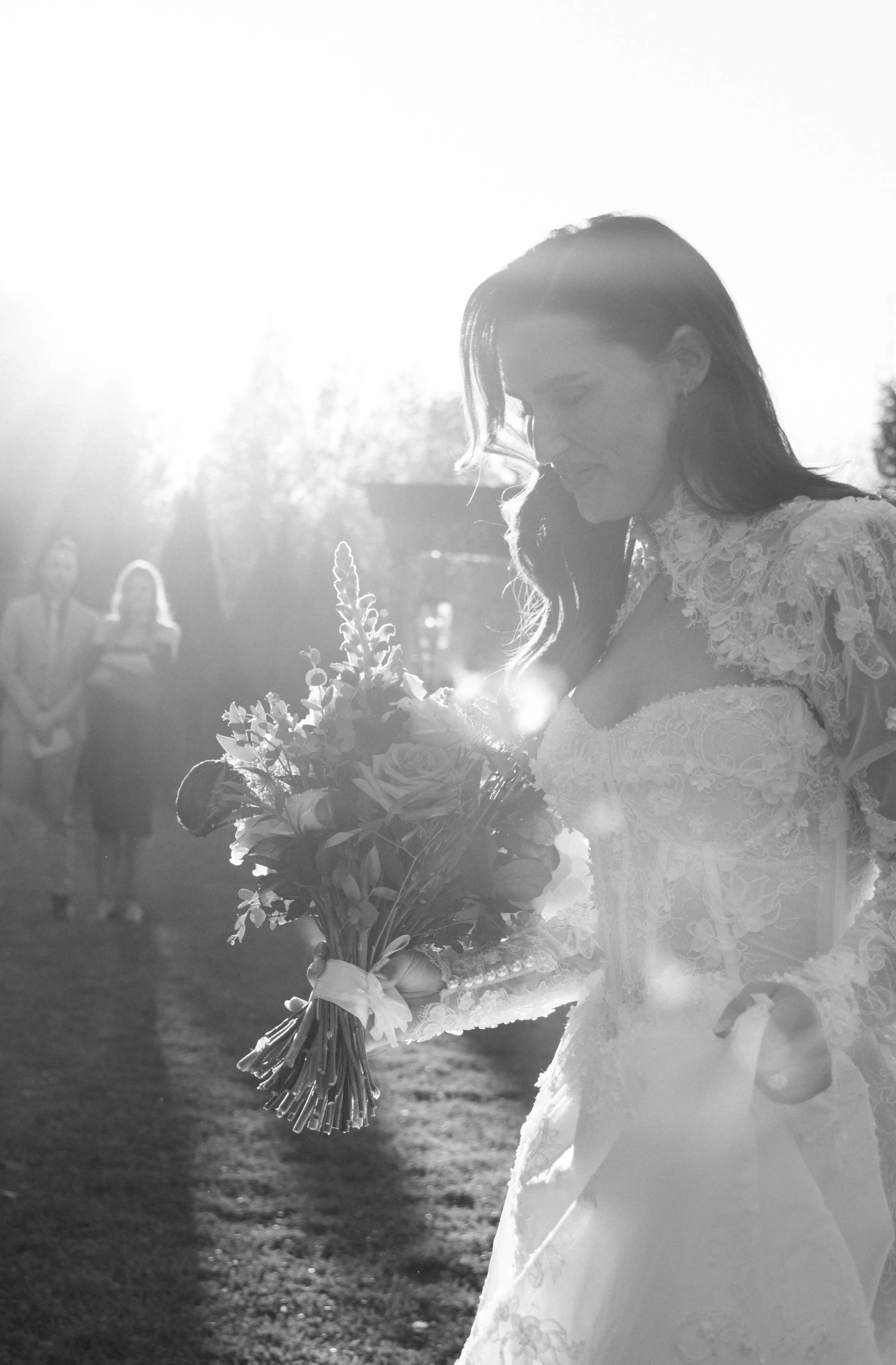 Black and white photo of a bride holding a bouquet of flowers, with sunlight shining from behind, and three people in the background.