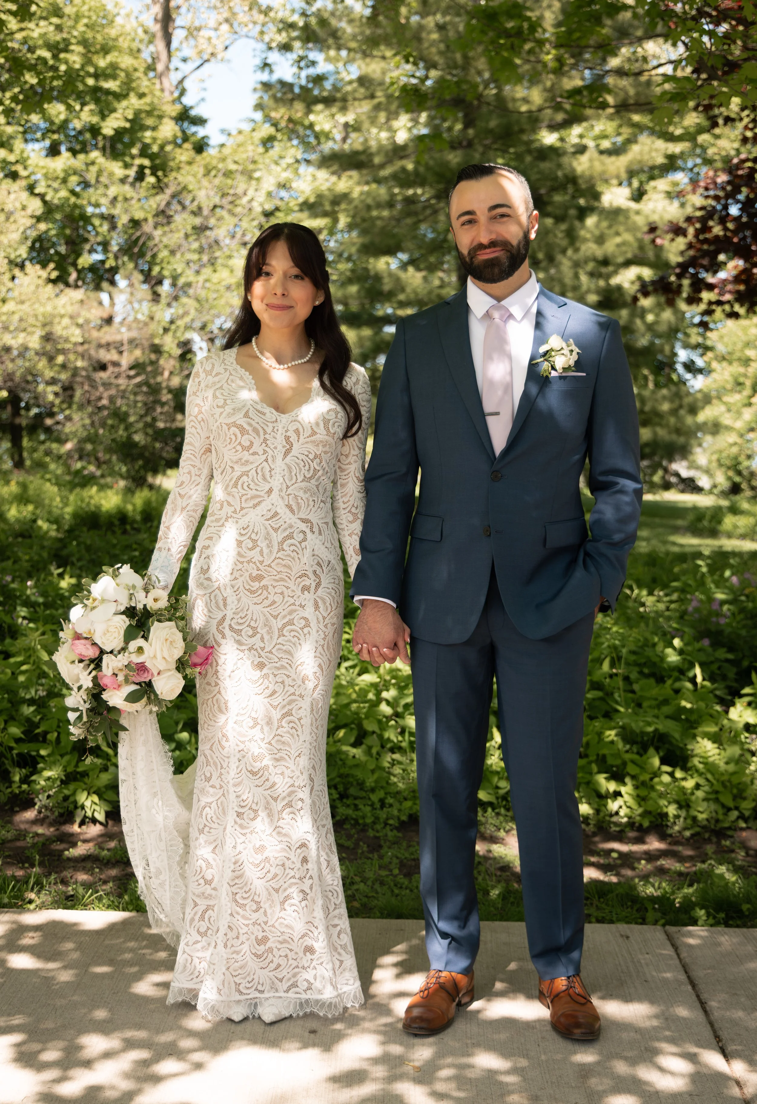 A bride and groom holding hands outdoors on a sunny day. The bride wears a long white lace dress and pearl necklace, holding a bouquet. The groom wears a blue suit with a pink tie and a flower boutonniere. They stand on a sidewalk with lush green tre