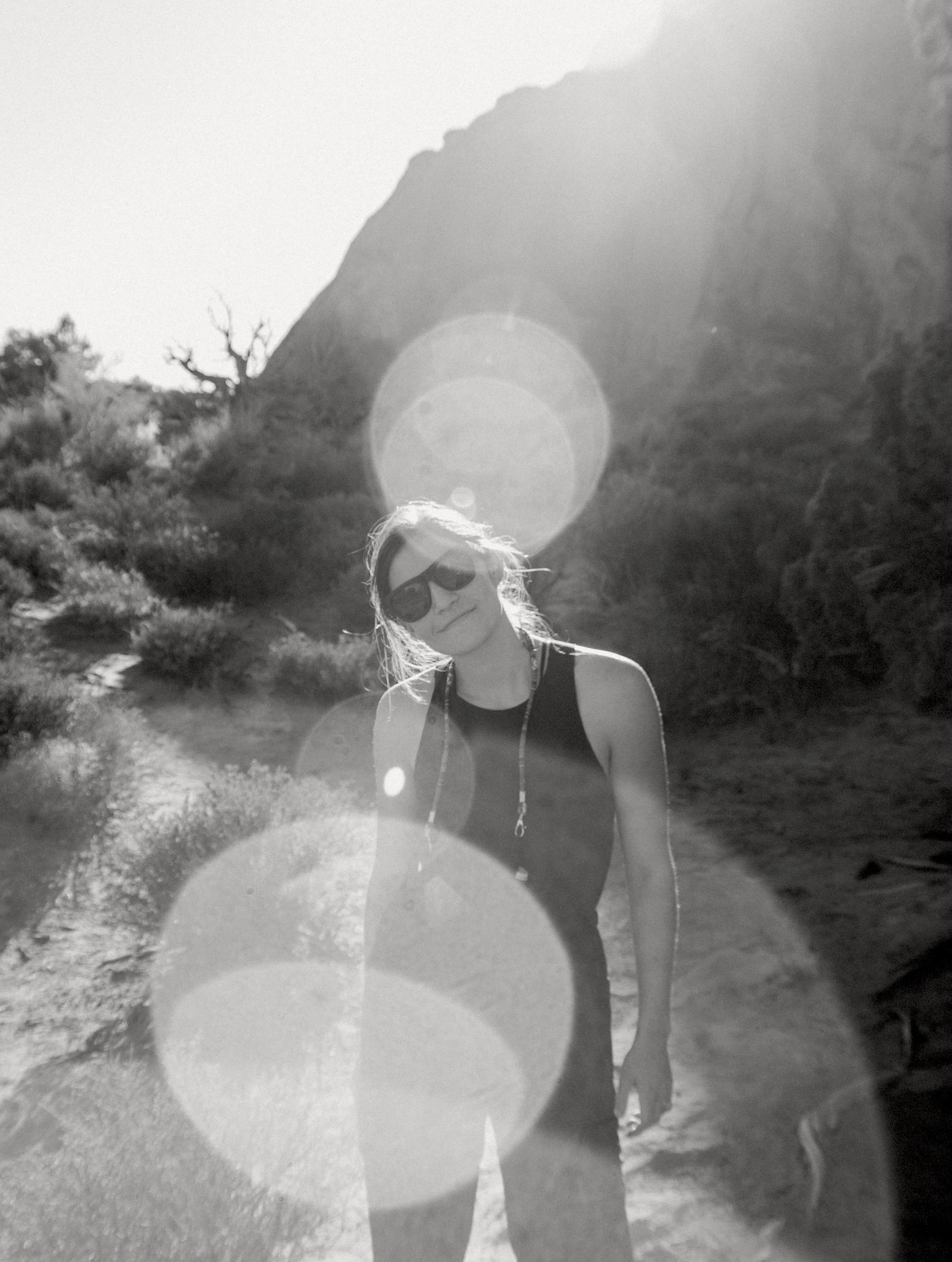 A woman with sunglasses and a sleeveless top standing outdoor with rocky terrain and hills in the background, sunset lighting creating lens flare effects.