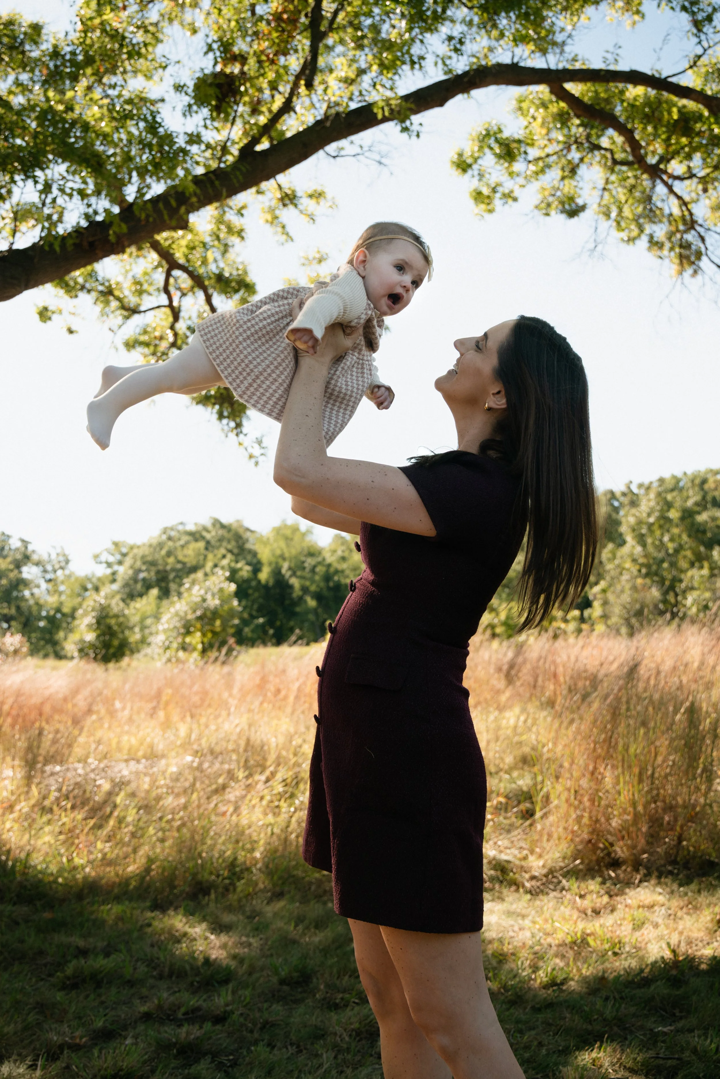 A woman with long dark hair lifting a young girl with blonde hair and a checkered dress into the air outdoors under a tree with green leaves, in a grassy field.